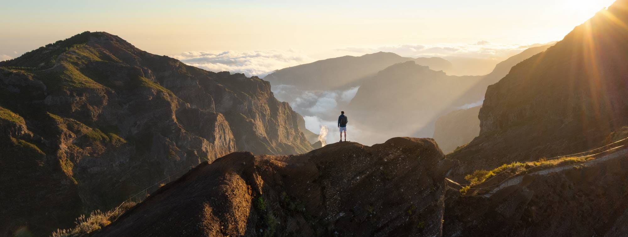 Personne debout sur une crête rocheuse au lever du soleil, face à une vallée montagneuse baignée de lumière.