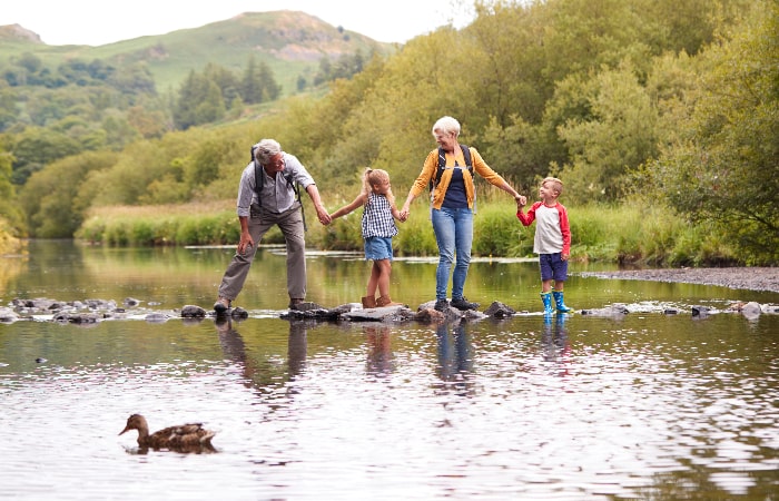 Famille marchant main dans la main sur des pierres dans une rivière, en pleine nature, avec un canard au premier plan.
